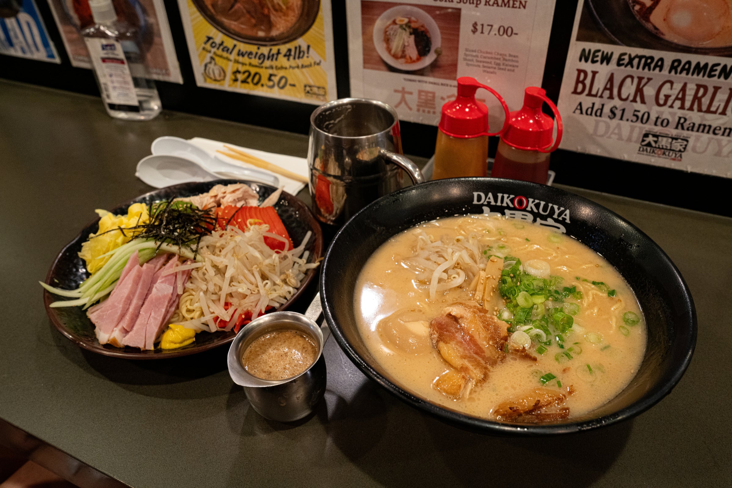 Bowl of ramen with toppings and side dishes on a table with promotional posters in the background.