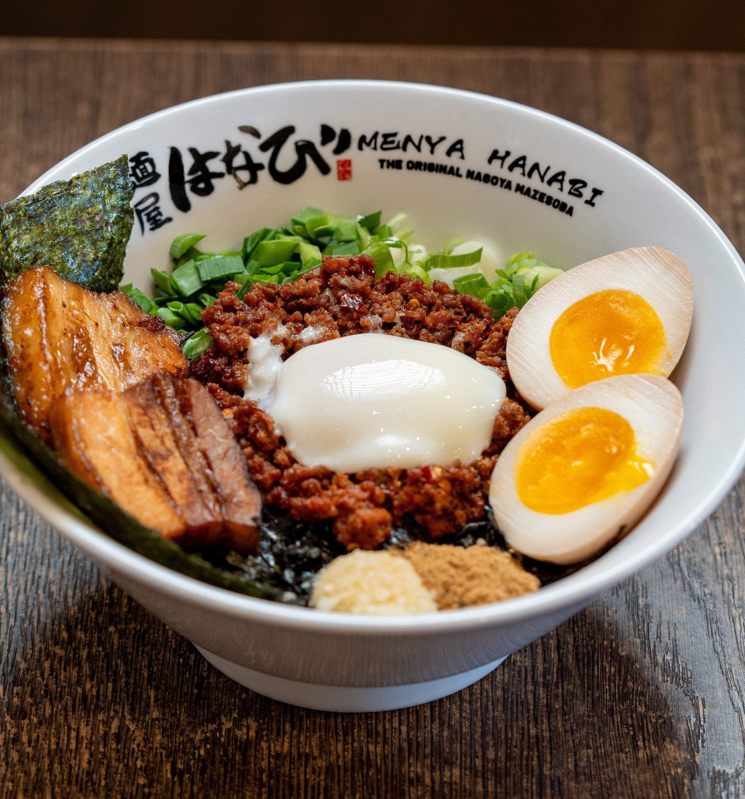 A bowl of mazesoba at menya haniabi in arcadia  ajitamama chashu minced pork nagaoya noodles japan authentic ramen on a dark wooden table, featuring a soft-boiled egg, sliced soft-boiled eggs, seaweed, greens, and braised pork.